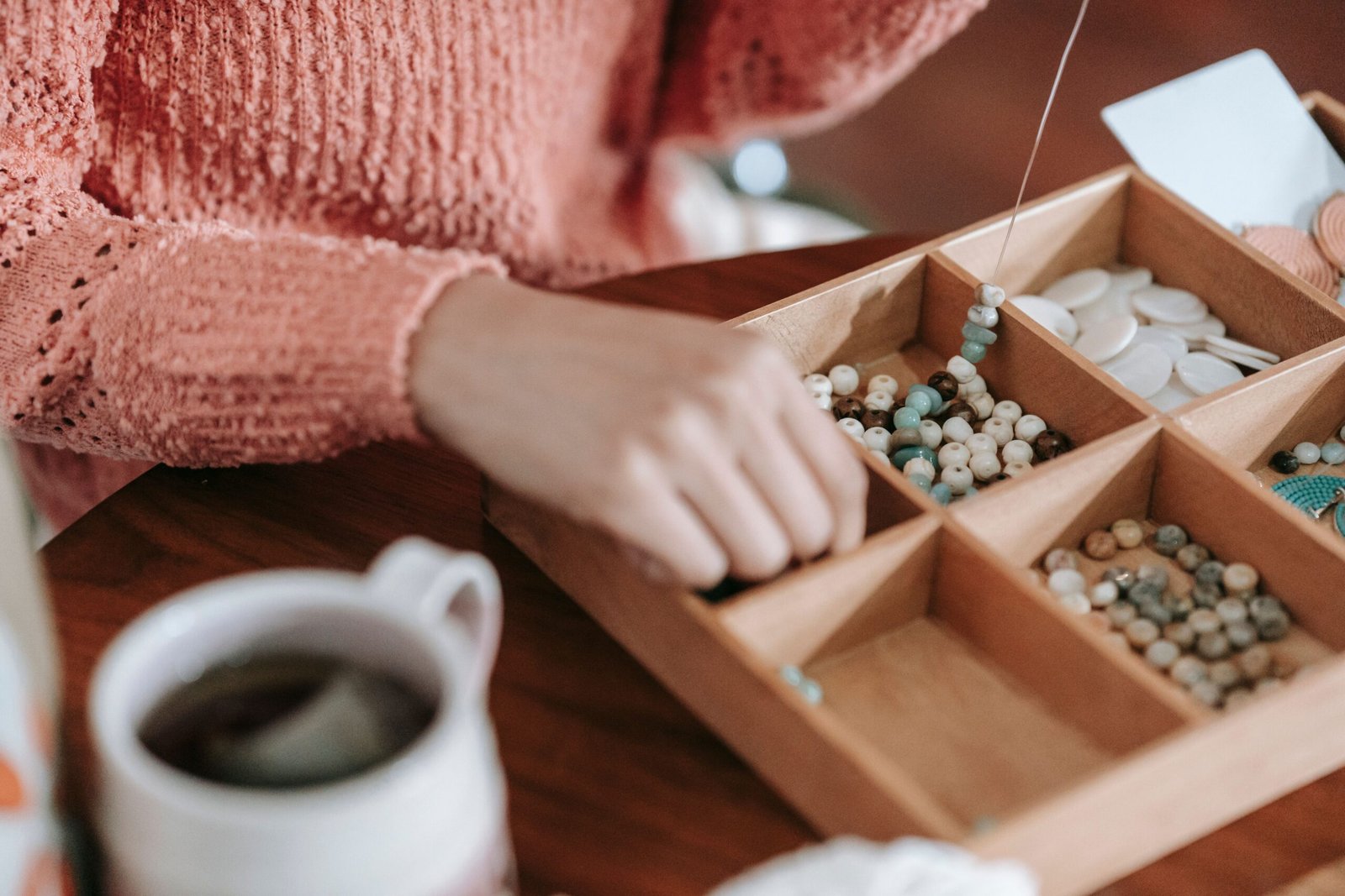 Crop unrecognizable female in casual outfit sitting at wooden table with cup of tea threading multicolored decorative beads and natural stones on fishing line while handiwork